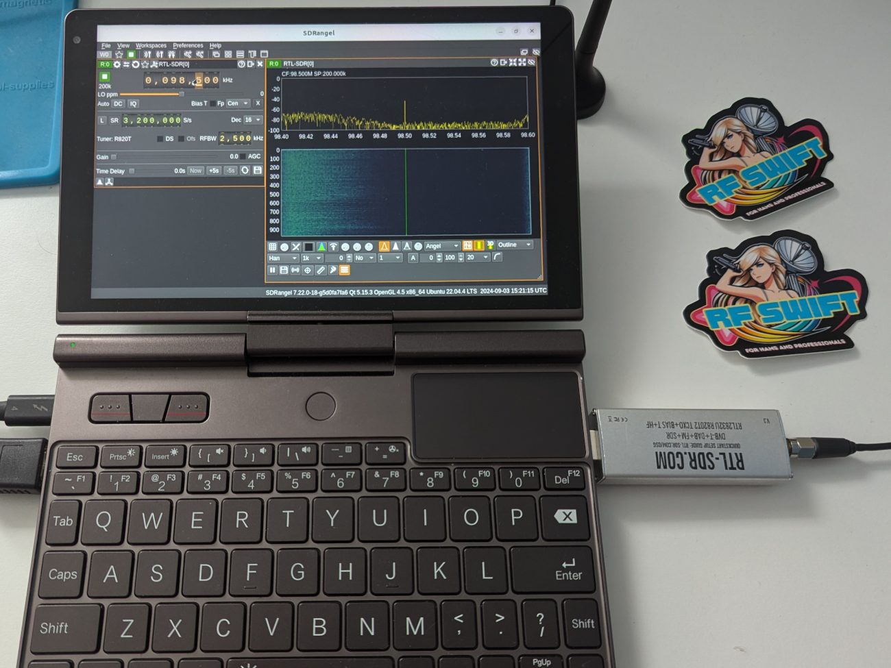 A GPD pocket laptop running SDRangel with an RTL-SDR dongle plugged in, showing an FM station at 98.5 MHz, next to two RF Swift stickers.